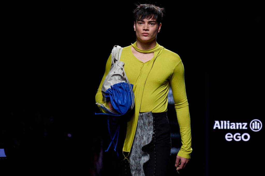 Un modelo masculino camina por la pasarela luciendo una camisa amarilla y un bolso azul.