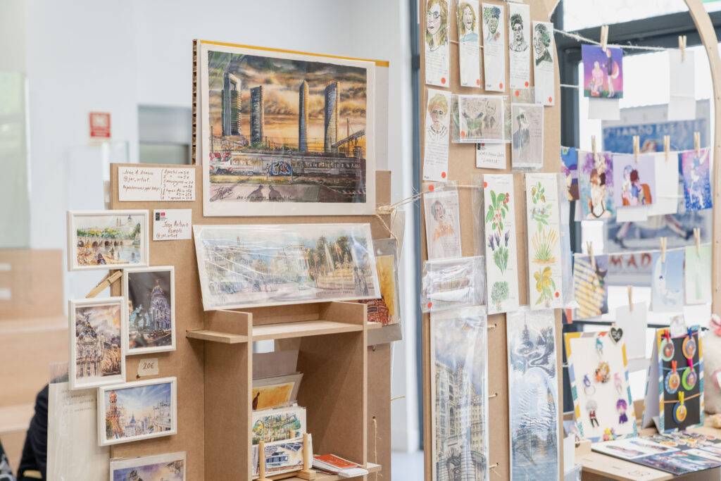 La imagen muestra una exposición de arte con varias obras enmarcadas y fotografías colgadas en una pared.