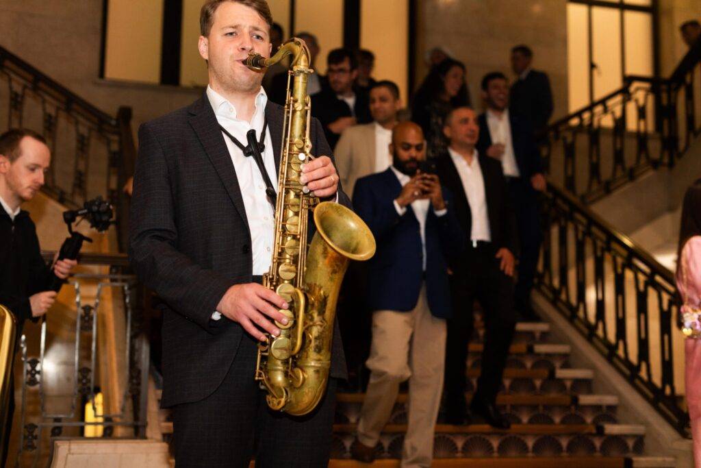 Un saxofonista toca en un evento mientras la gente se mueve por el fondo.