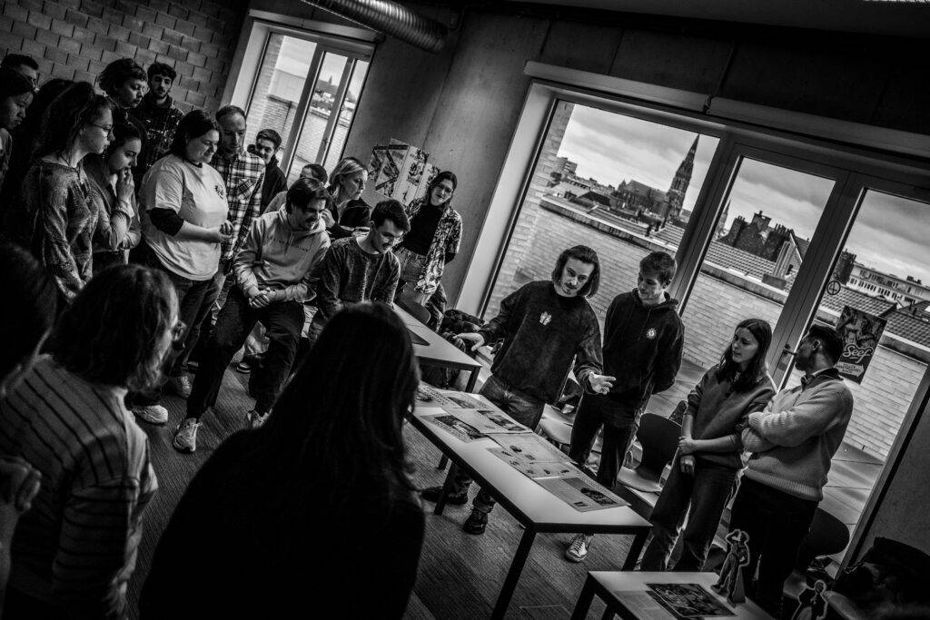 Un grupo de personas observa mientras alguien presenta documentos en una sala con grandes ventanas.