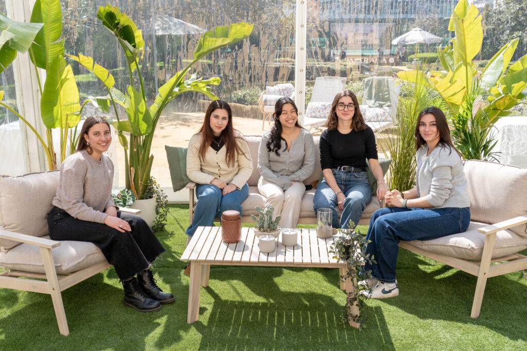 Cinco mujeres se reúnen sonrientes en un ambiente acogedor rodeadas de plantas.