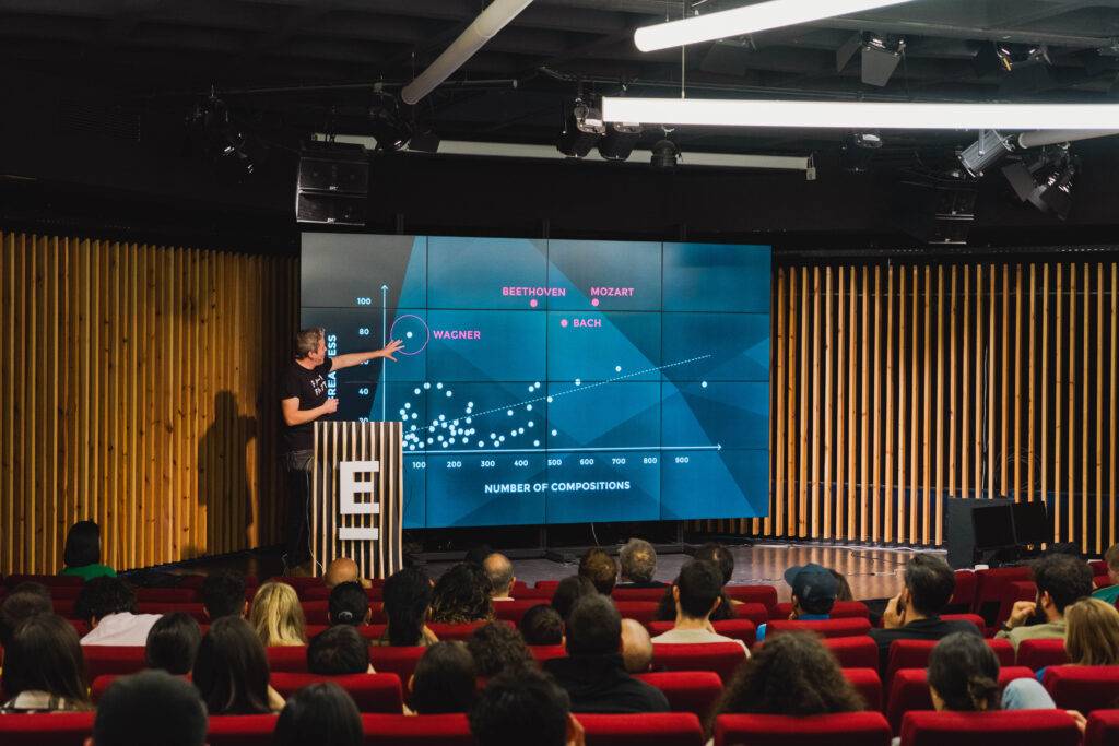 Un presentador muestra un gráfico sobre la composición musical en una sala llena de público.