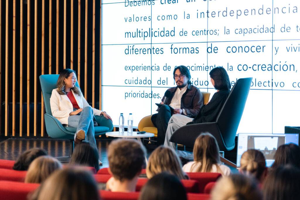 Una charla en un auditorio con tres ponentes discutiendo sobre la co-creación y la interdependencia en el aprendizaje.