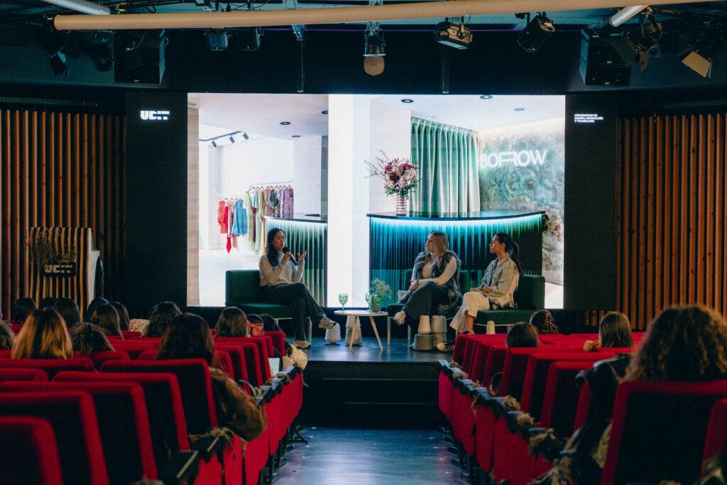 Una charla en un auditorio con un fondo de pantallas que muestran ropa y asistentes en sillones rojos.