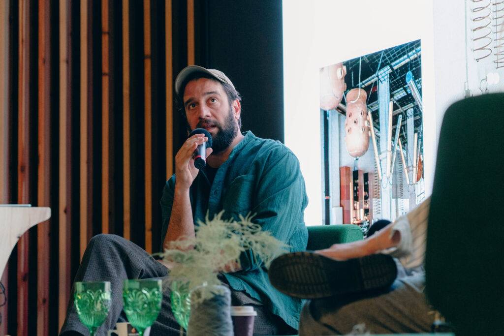 Un hombre con barba y gorra sostiene un micrófono mientras habla en un evento.