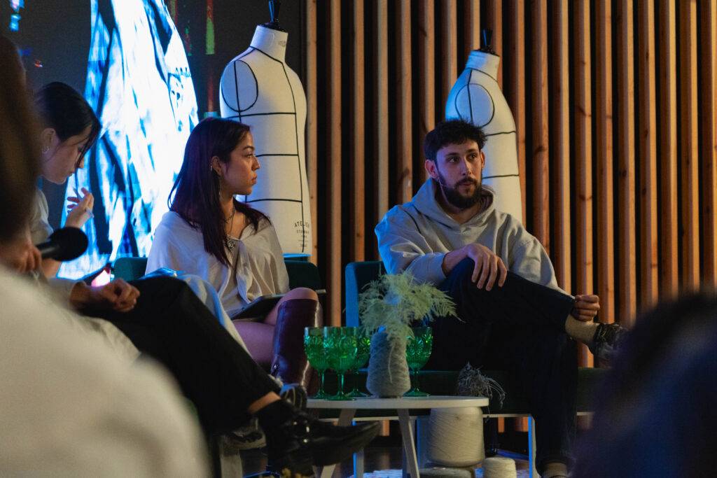 Un panel de discusión con un grupo de personas sentadas, una de ellas viste un suéter gris y otra lleva una blusa blanca.