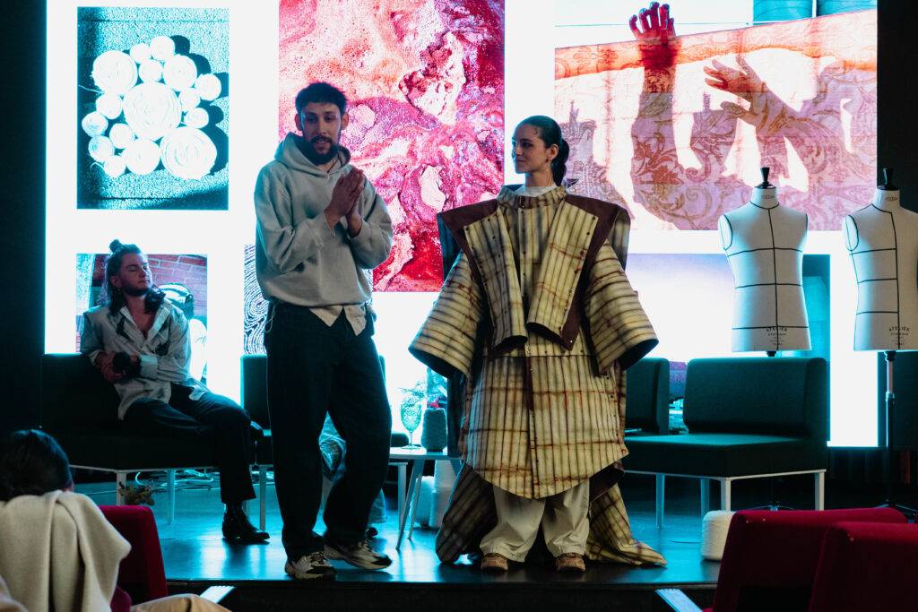 Un grupo de personas en una pasarela, con un modelo vistiendo una atuendo llamativo mientras otro individuo habla.