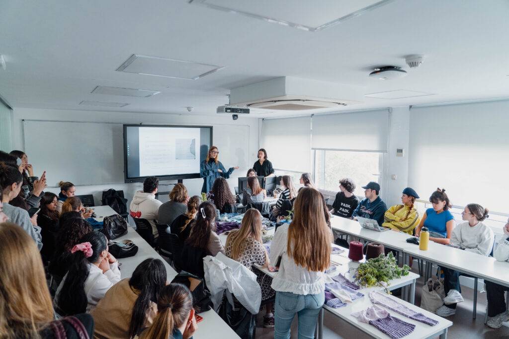 Una clase en un aula donde dos presentadoras están hablando frente a una audiencia de estudiantes atentos.