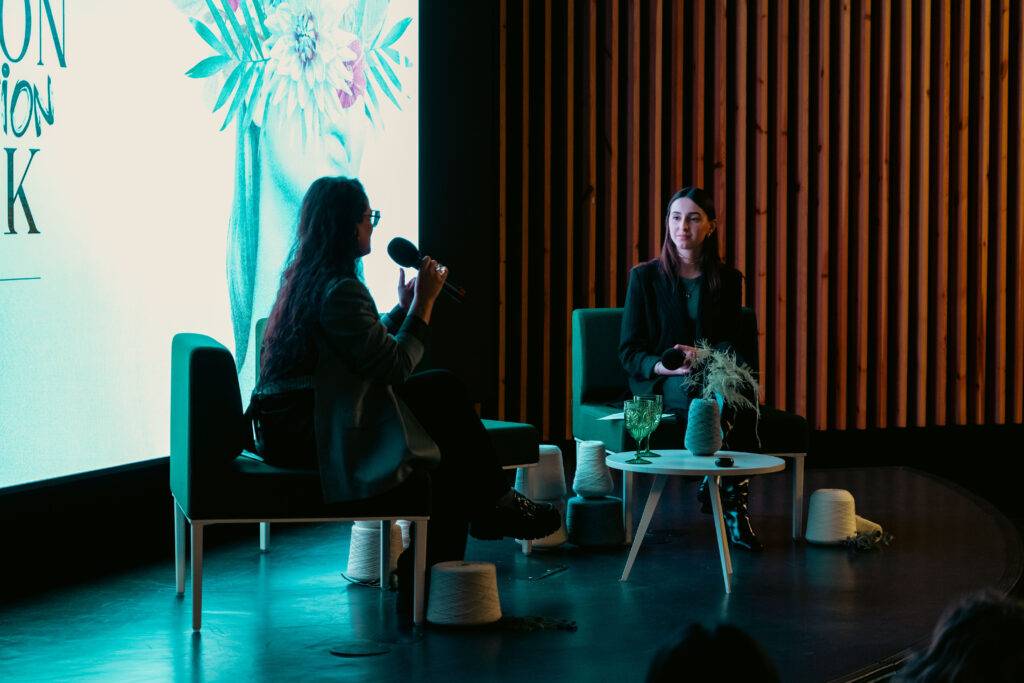 Dos mujeres conversan en un evento sobre moda en un escenario minimalista.