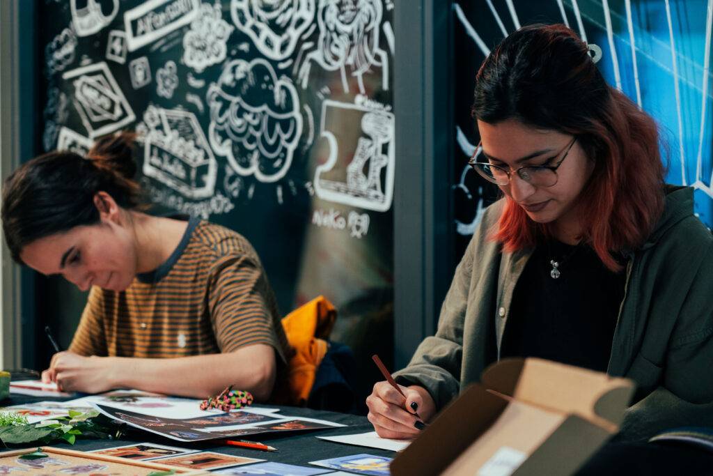 Dos personas se encuentran dibujando y pintando en una mesa con un fondo decorado con dibujos.