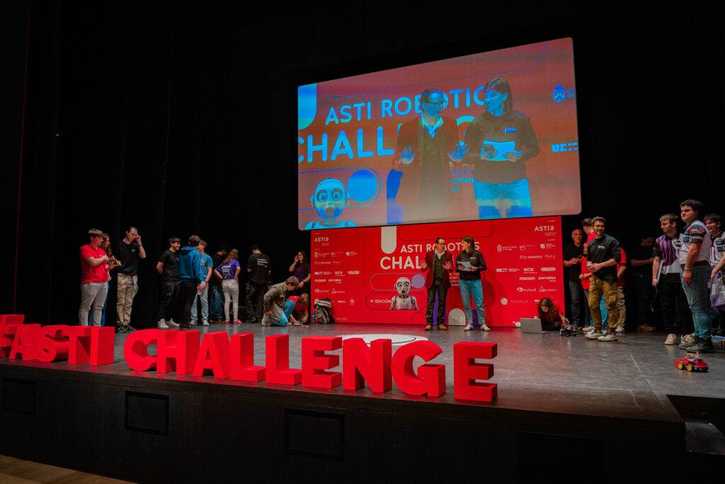 La imagen muestra un evento en un escenario con participantes y una presentación en pantalla sobre el Asti Robotics Challenge.