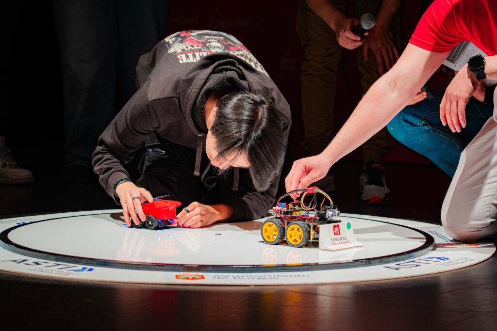 Un grupo de personas observa a un participante que ajusta un robot en una competición sobre una superficie circular.
