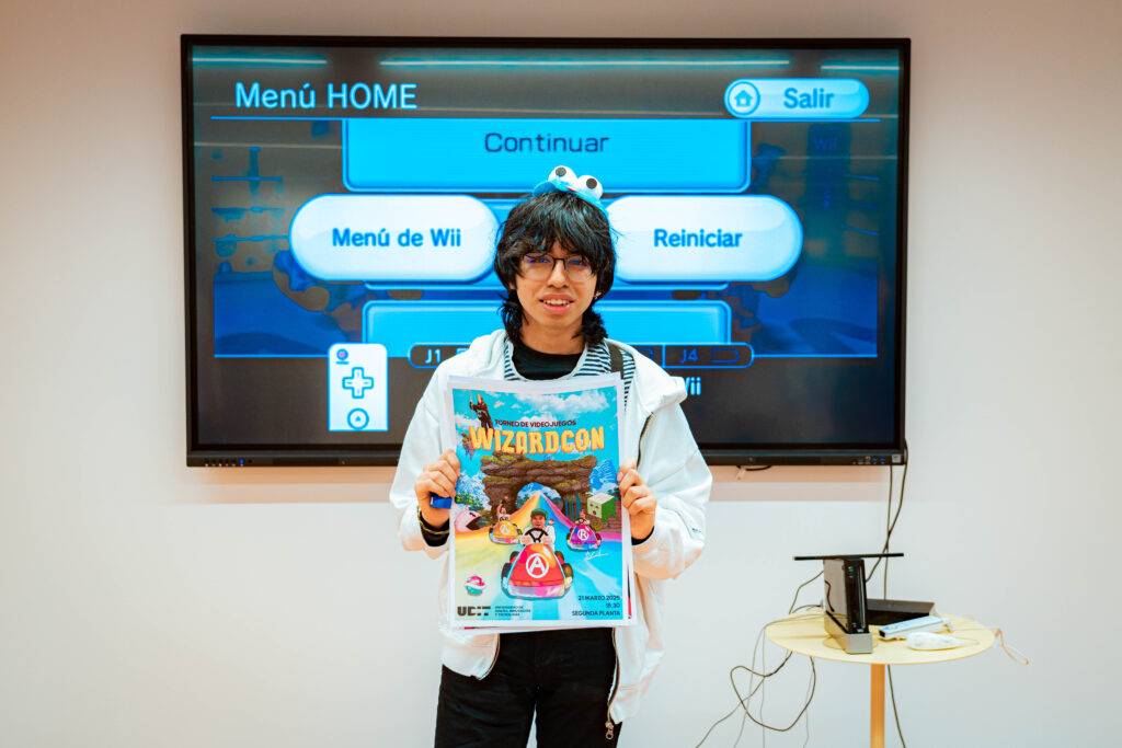 Un joven sostiene un cartel de un evento llamado Wizardcon frente a una pantalla que muestra el menú de una consola de videojuegos.