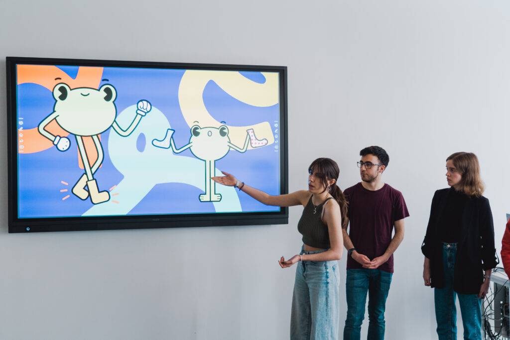 Cuatro personas observan una pantalla que muestra ilustraciones de ranas animadas.