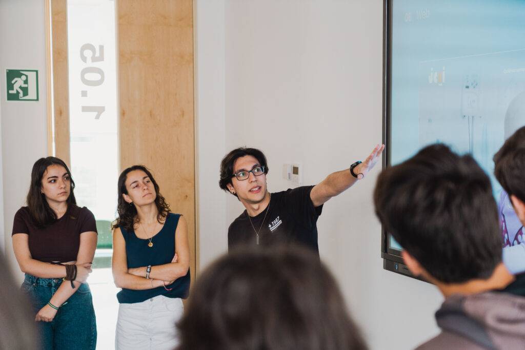 Un grupo de personas escucha atentamente a un joven que está explicando algo frente a una pantalla.