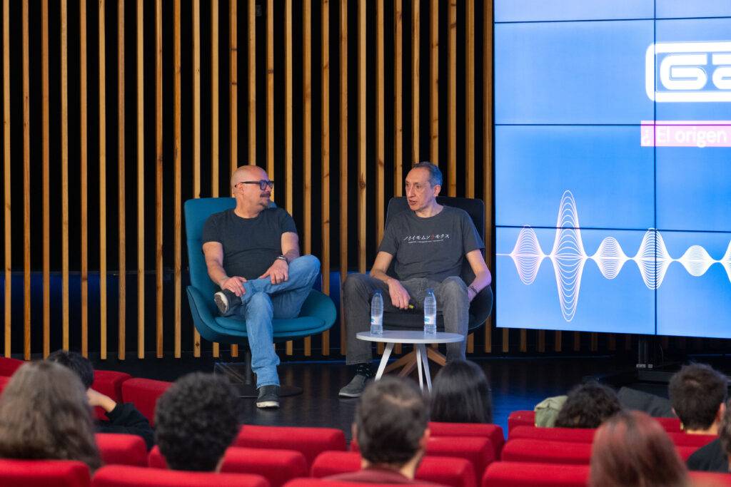 Una conferencia en la que dos oradores discuten sobre un tema frente a un público sentado en sillones rojos.