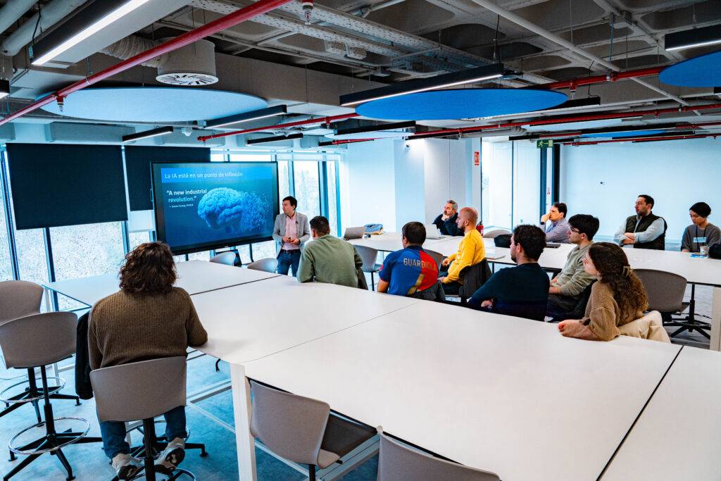 Una reunión en un espacio moderno donde un ponente presenta información a un grupo de personas sentadas alrededor de una mesa.