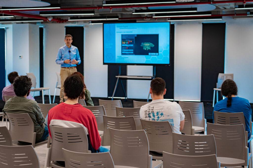 Se muestra a una persona presentando frente a una audiencia en una sala moderna con un proyector.
