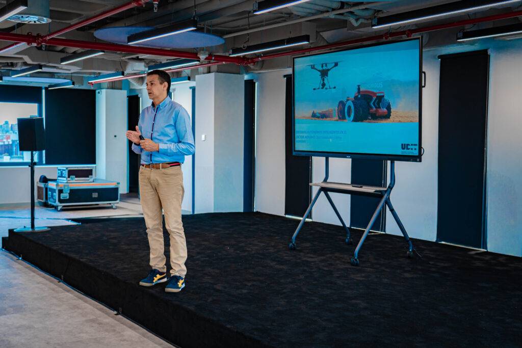 Un hombre presenta en un escenario mientras se muestra una imagen de un tractor y un dron en una pantalla grande.