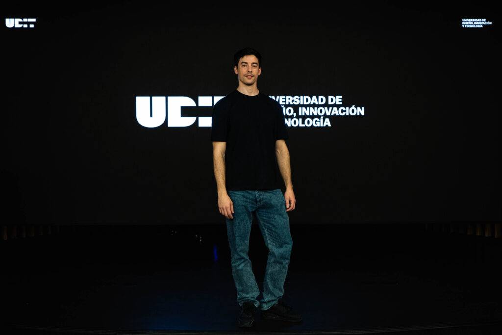 Un hombre de pie frente a una pantalla con el logotipo de una universidad.