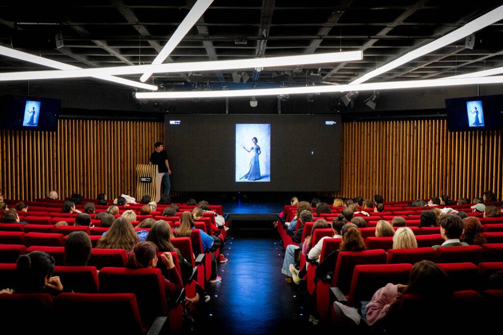 Una presentación en un auditorio donde una persona habla frente a una gran pantalla mostrando una imagen de una mujer.