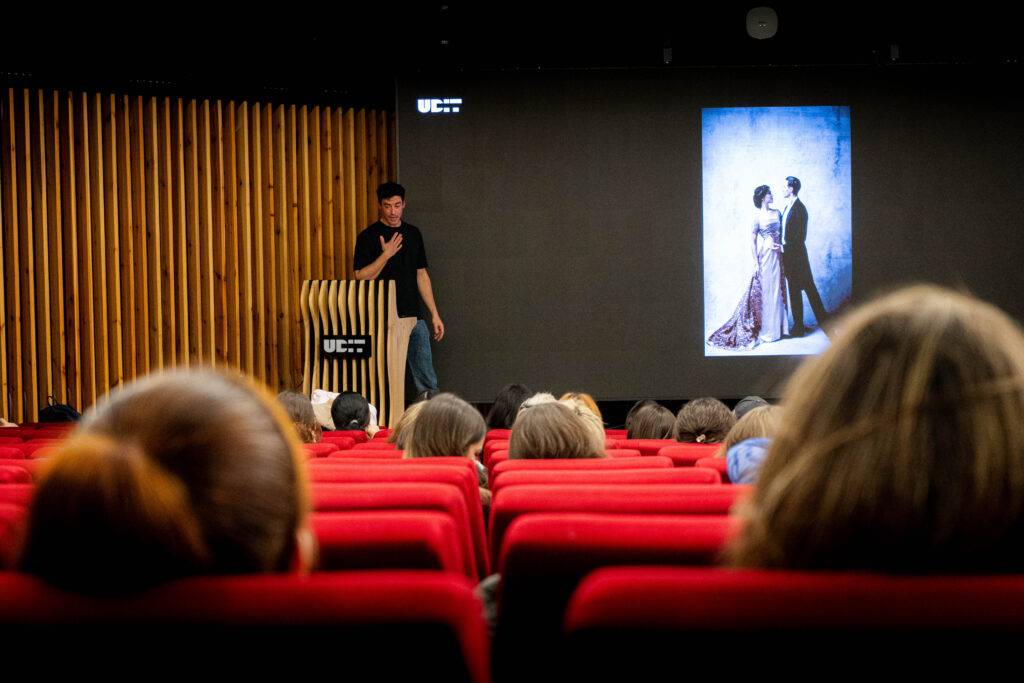 Un conferencista presenta una imagen en una pantalla mientras la audiencia lo observa desde sus asientos en un auditorio.