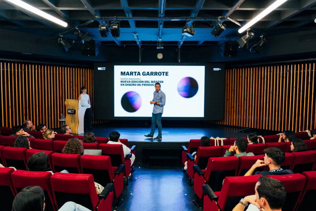 Un hombre presenta en un auditorio mientras una mujer escucha, con una pantalla que muestra información sobre Marta Garrote.