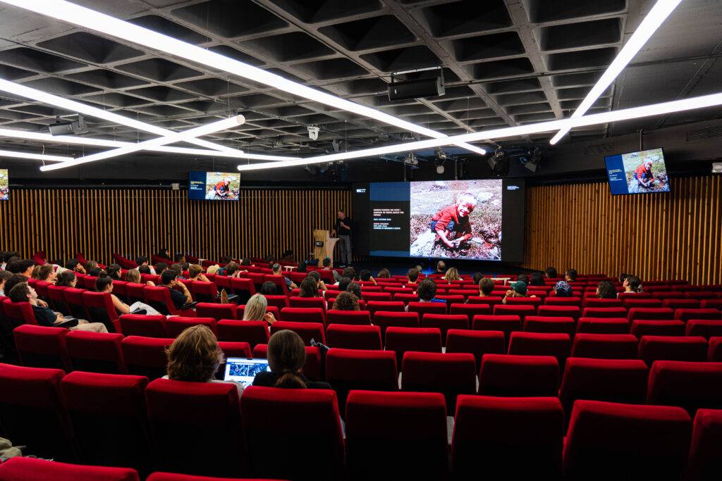 Una sala de conferencias con varias personas sentadas y un ponente en el escenario proyectando una imagen.