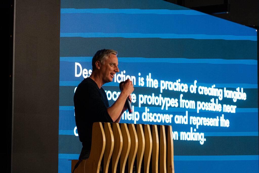 Un hombre está hablando en un evento con un micrófono frente a una pantalla que muestra texto sobre diseño.