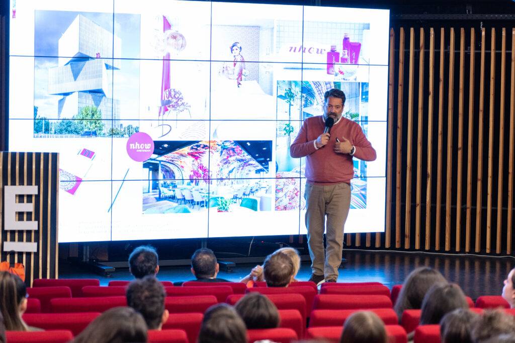 Una persona habla en una conferencia frente a una pantalla grande mostrando imágenes coloridas.