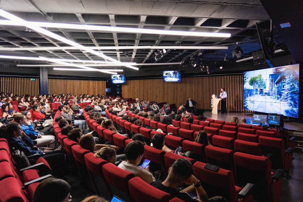 Una sala de conferencias llena de público, con pantallas y un ponente en el escenario.