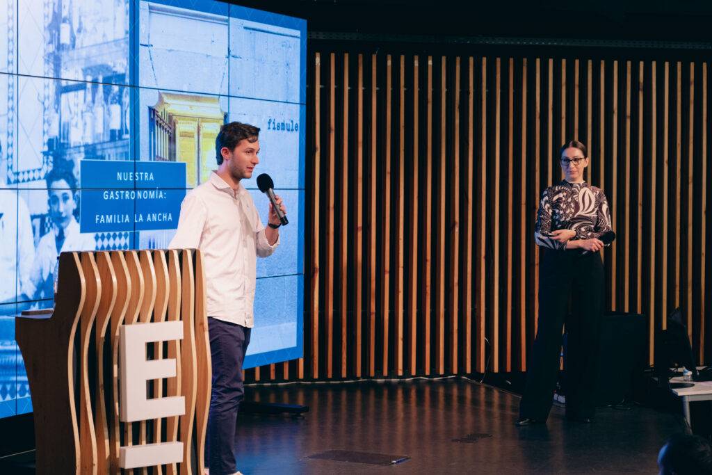 Un hombre habla en un escenario mientras una mujer lo observa, en un evento de presentación.