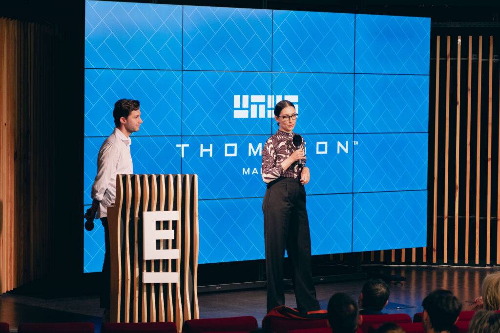 Un presentador y una presentadora están hablando frente a una gran pantalla en un evento.