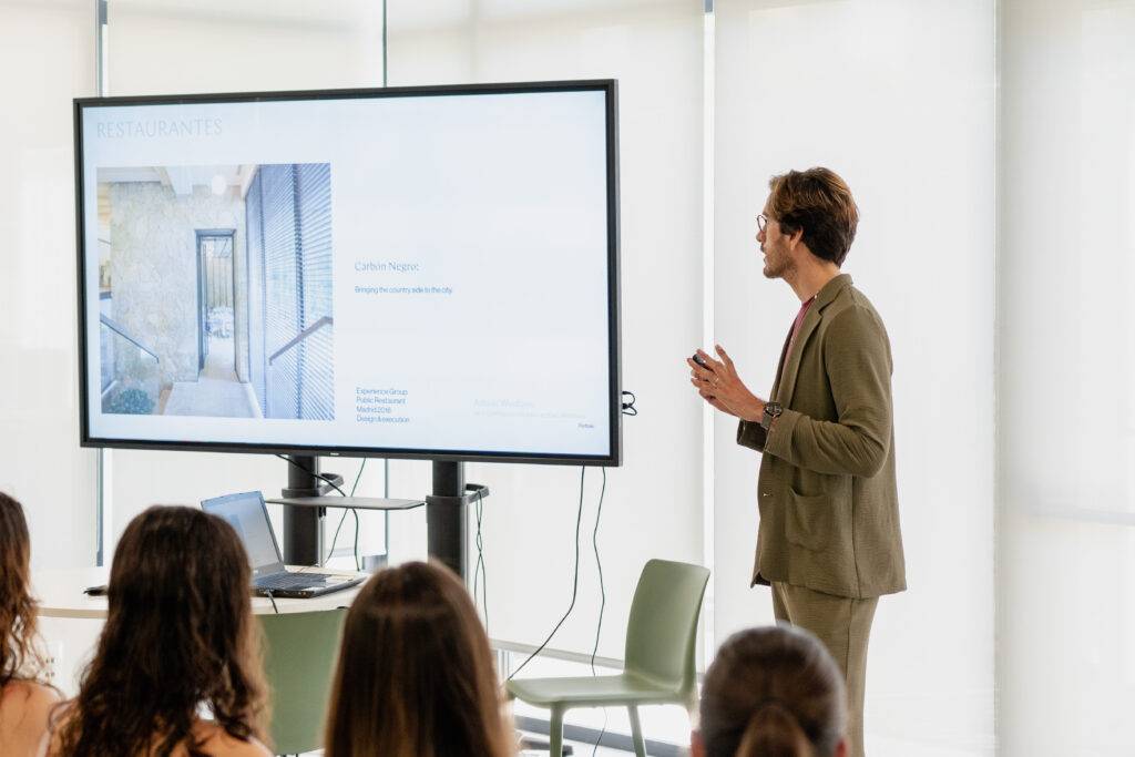 Un presentador habla sobre un proyecto arquitectónico frente a una audiencia en una sala iluminada.
