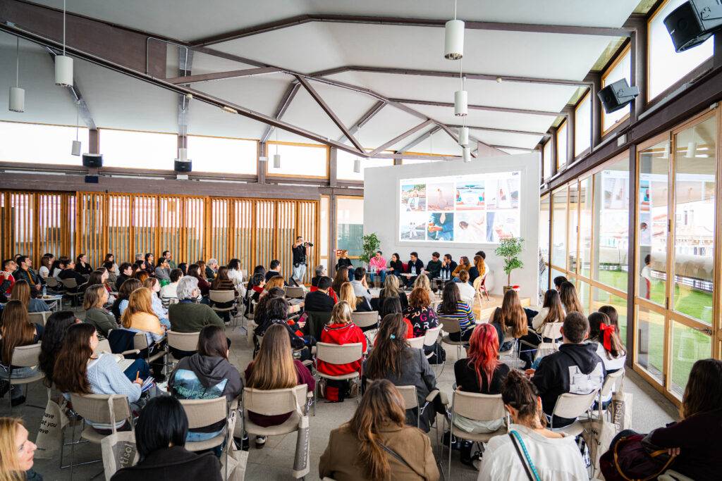 Un grupo de personas asiste a una charla en un salón con grandes ventanales y una pantalla al fondo.