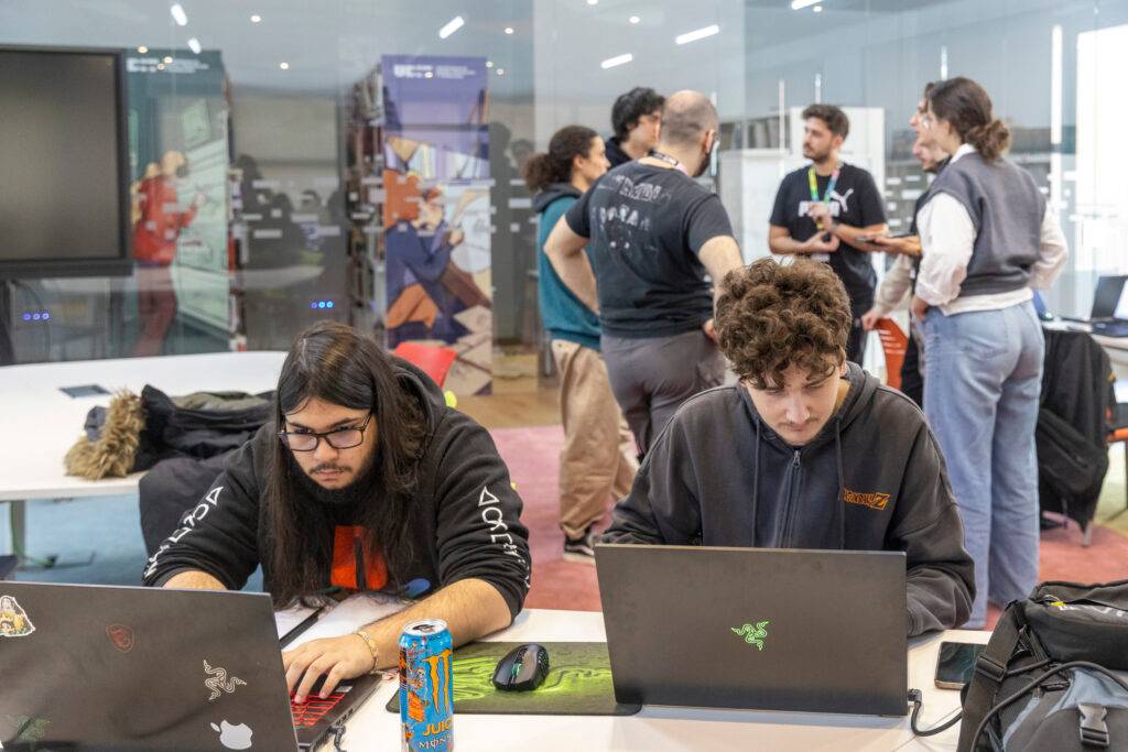 Dos personas trabajan en sus portátiles mientras un grupo de personas conversan al fondo en un ambiente de oficina.