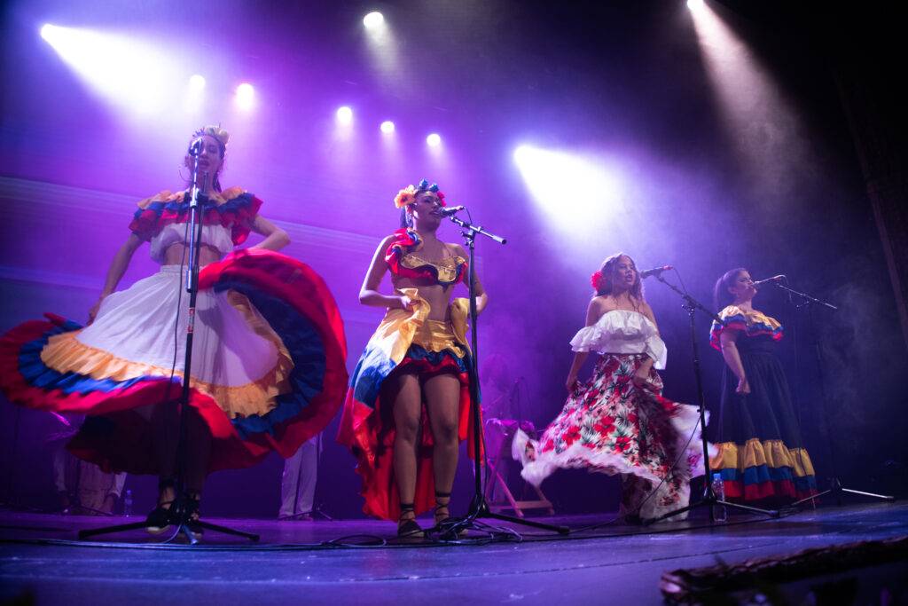 Cuatro mujeres vestidas con trajes tradicionales realizan una presentación en un escenario iluminado.