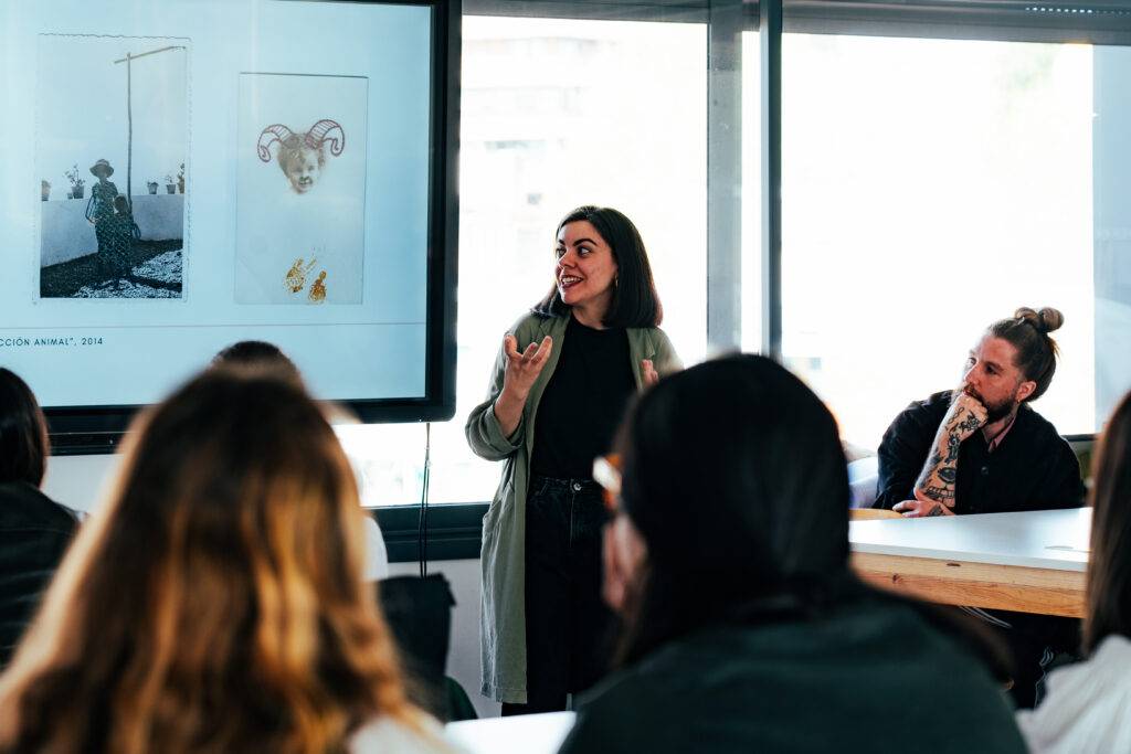 Una mujer está presentando en una sala frente a un grupo de personas, mostrando imágenes en una pantalla.