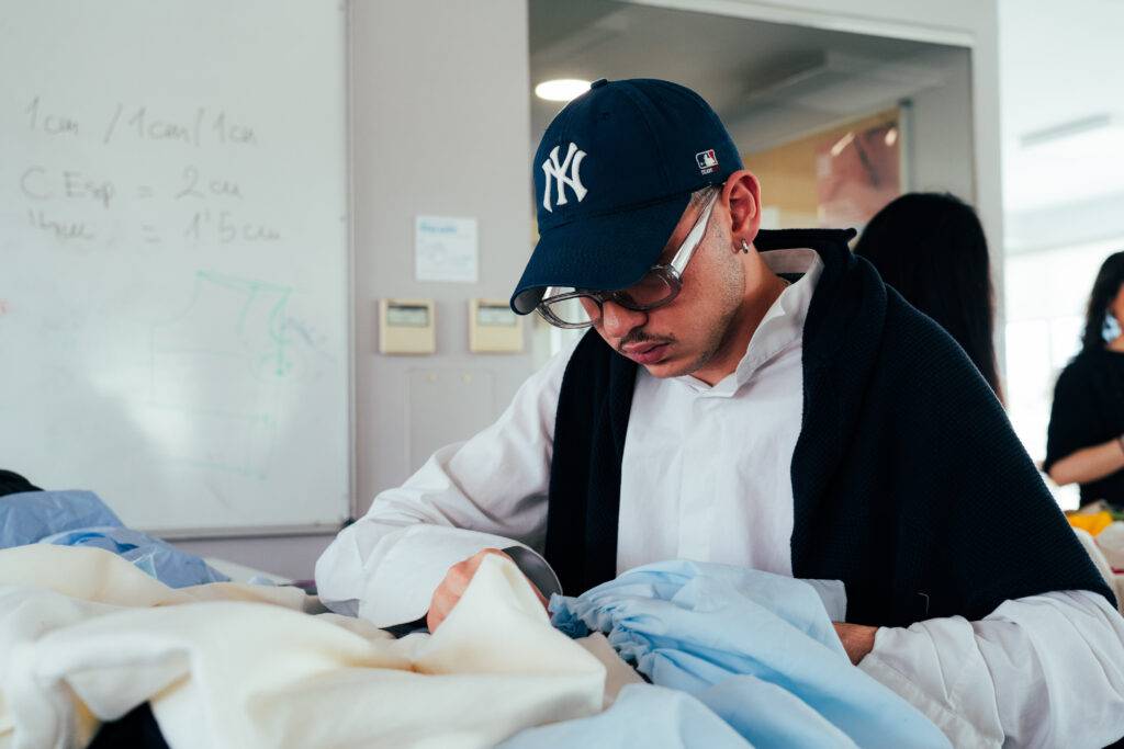 Un joven está trabajando en una mesa con varias prendas de ropa en un ambiente educativo.