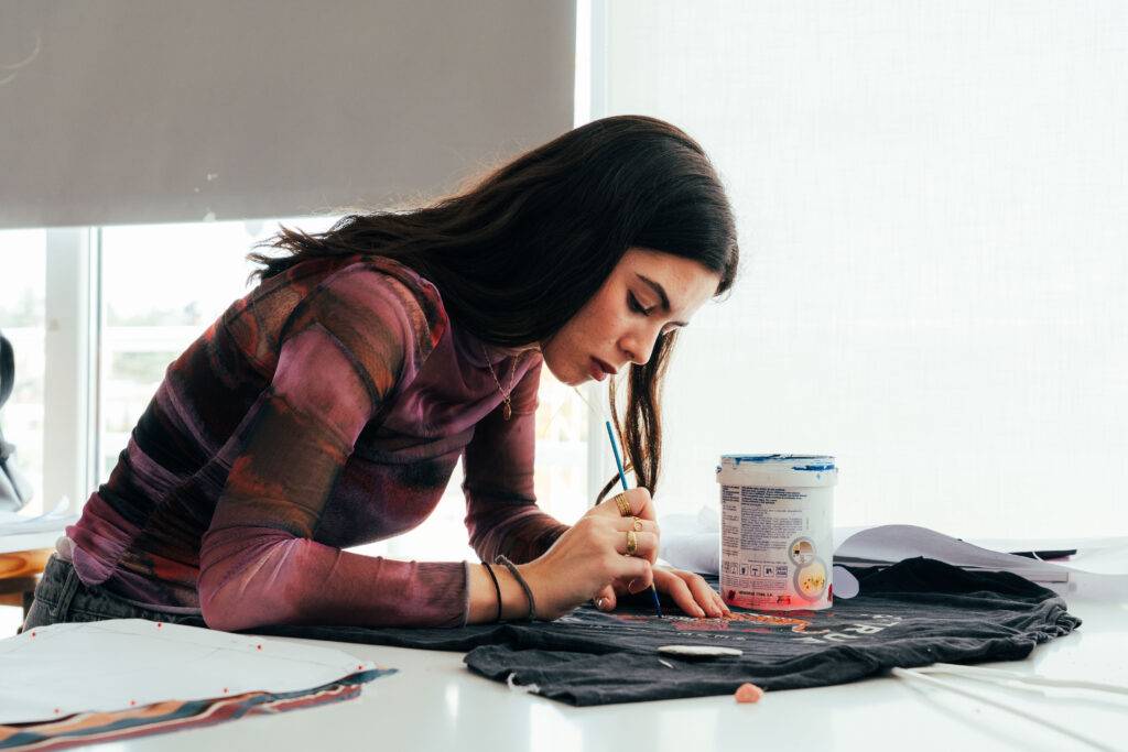 Una joven artista está pintando con atención sobre una camiseta negra en un ambiente bien iluminado.