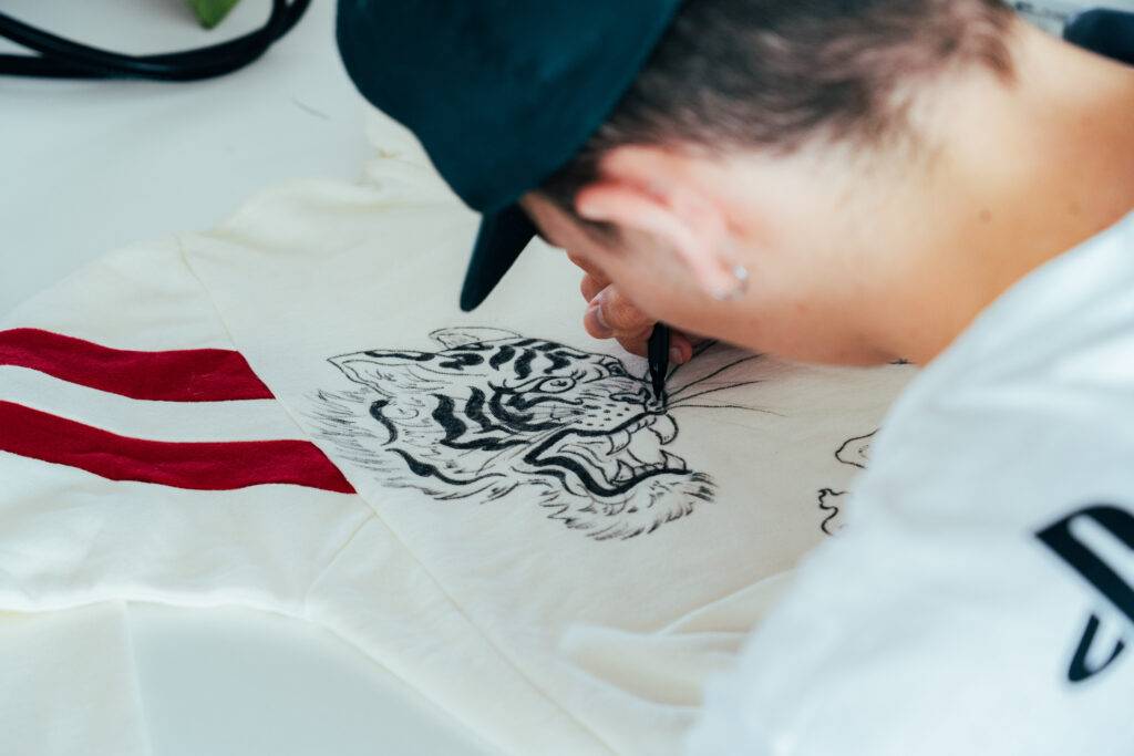 Un joven dibuja un tigre sobre una camiseta blanca con rayas rojas.