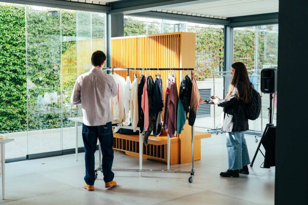 Una pareja de personas observa y revisa ropa en un escaparate moderno con una pared de cristal y vegetación al fondo.