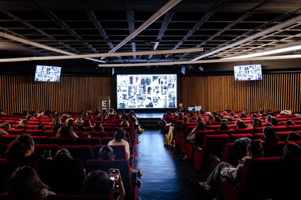 Una sala de cine llena de personas sentadas en butacas rojas, mirando una pantalla grande con imágenes.