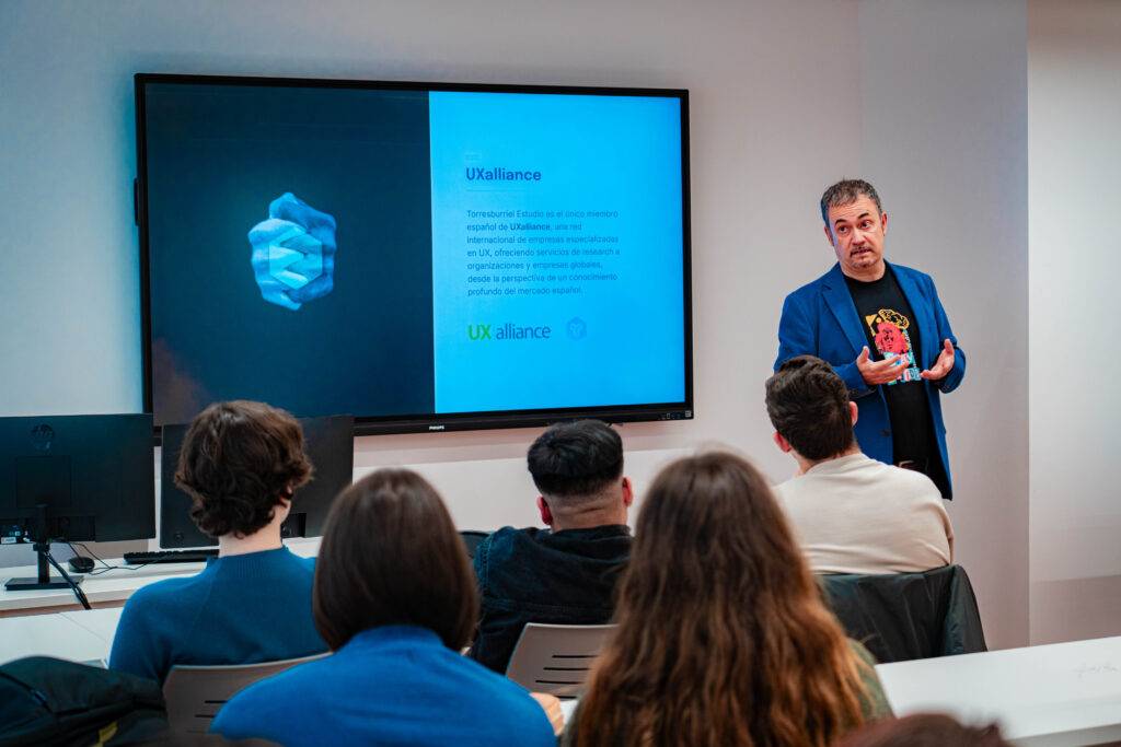 Un presentador habla frente a un grupo de personas en un aula moderna mientras se muestra una diapositiva sobre UX alliance.