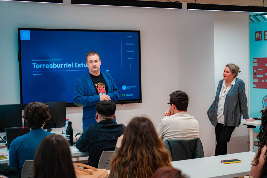 Una presentación en un aula donde un hombre está hablando frente a un grupo de estudiantes y una mujer lo está observando.