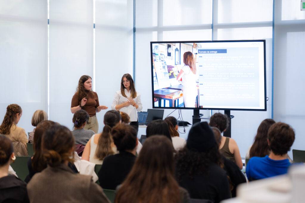 Dos mujeres están presentando frente a un público en un aula con una pantalla grande.