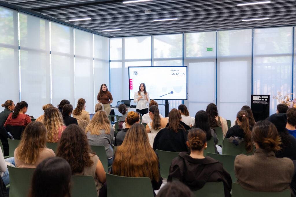 Una conferencia en un aula con varias personas asistentes, donde dos mujeres están presentando un tema en la pantalla.
