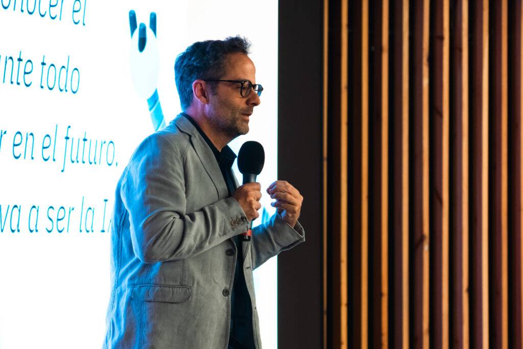 Un hombre habla en un escenario mientras sostiene un micrófono y se proyecta una diapositiva de fondo.