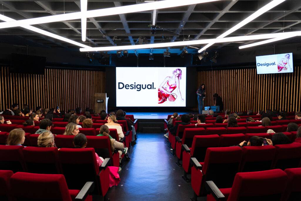 Una sala de conferencias con asientos rojos y una pantalla que muestra el logo de Desigual.