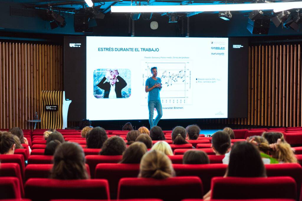 Un conferencista presenta sobre el estrés laboral en un auditorio con asientos rojos y una pantalla detrás.